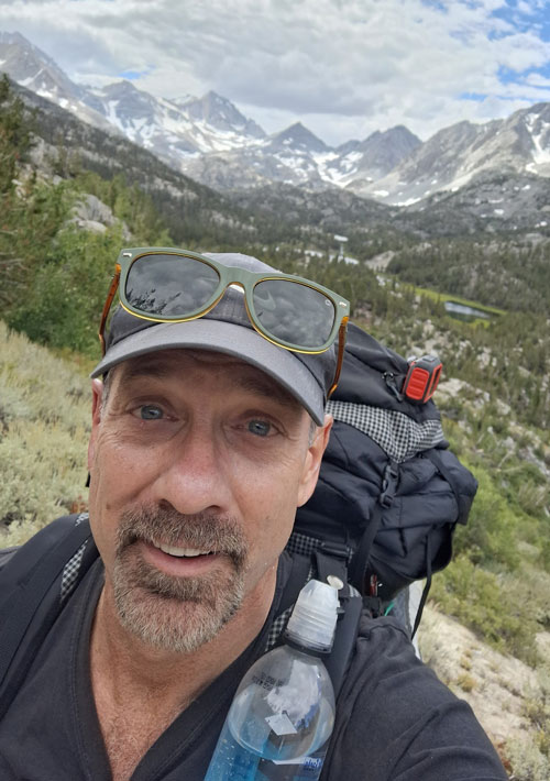Andrew Heyl smiling while hiking with scenic mountains behind him